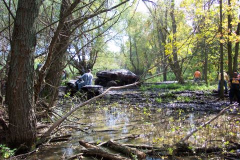1978 Ford Bronco - An off-road vehicle is stuck in a muddy, wooded area with several people nearby.