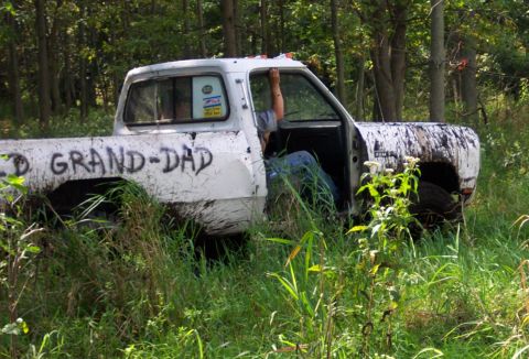 1978 Ford Bronco - A weathered white pickup truck sits abandoned in a wooded area, its side bearing a hand-painted message.