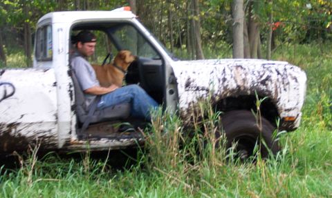 1978 Ford Bronco - A man and his dog sit in a mud-covered white pickup truck parked in a grassy, wooded area.