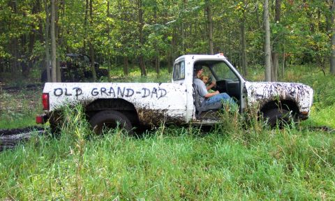 1978 Ford Bronco - A white pickup truck, labeled "OLD GRAND-DAD", is stuck in a muddy, grassy area with trees in the background. Another vehicle is partially…
