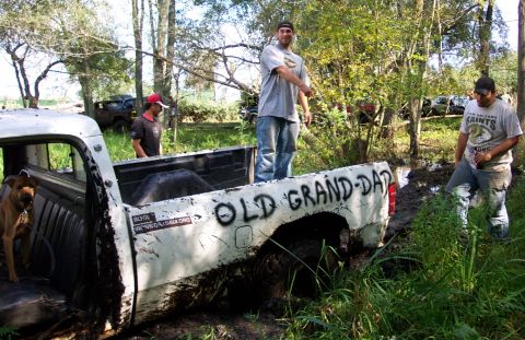 1978 Ford Bronco - A white pickup truck, spray-painted with 'OLD GRAND-DAD', is stuck in the mud with several people nearby.