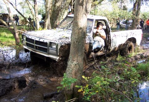1978 Ford Bronco - A white Dodge pickup truck is stuck deep in mud and trees, with other vehicles visible in the background.