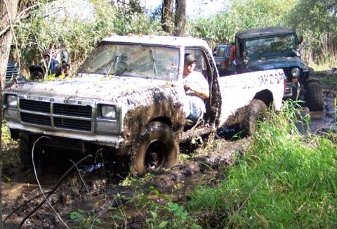 1978 Ford Bronco - A white Dodge pickup truck is stuck deep in the mud, with a driver visible inside the cab.