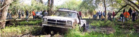 1978 Ford Bronco - A group of people watch as a white pickup truck struggles in a muddy, wooded area.