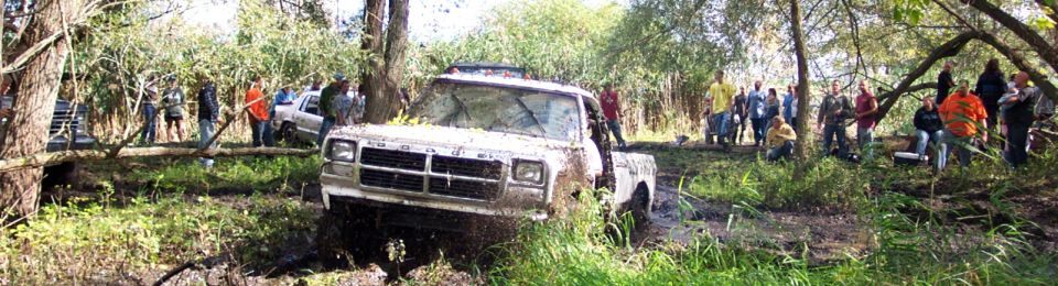 Mud-splattered white pickup truck stuck in a muddy ditch with onlookers