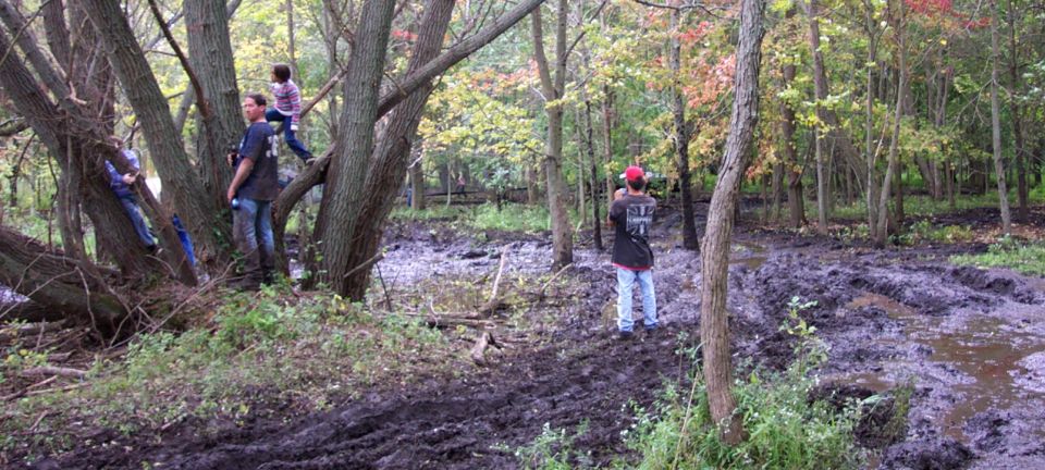 People in a muddy wooded area with trees and fallen branches