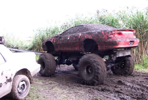 1978 Ford Bronco - A red monster truck is heavily covered in mud and appears stuck in a muddy area, with another vehicle attempting to tow it.
