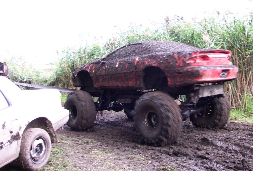 Red monster truck stuck in mud, being pulled by a white vehicle
