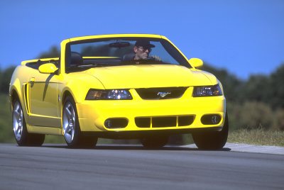 2003 Ford Mustang - A bright yellow 2003 Ford Mustang convertible is captured in a three-quarter front view, driving on a paved road under a clear blue sky.