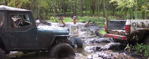 1978 Ford Bronco - Two off-road vehicles are stuck in a muddy forest area with a man posing between them.