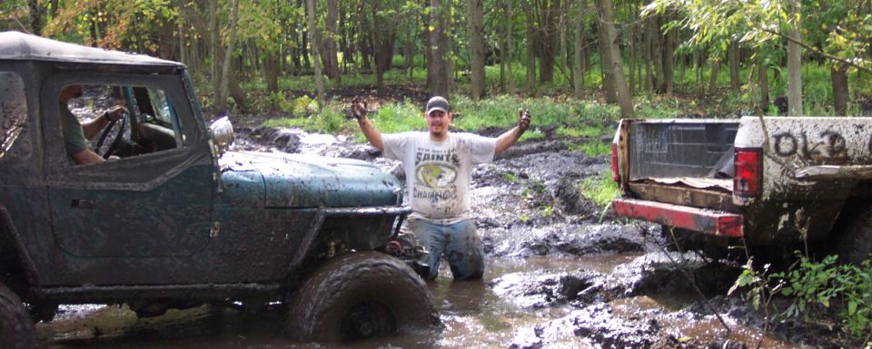 Muddy Jeep and truck in woods with guy in middle of mud pit