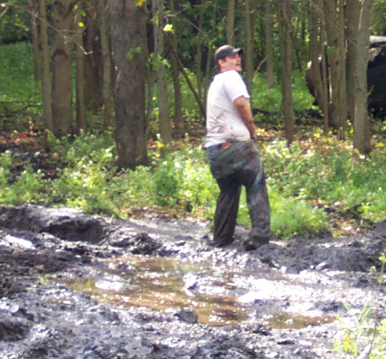 Man in dirty jeans and white t-shirt stands in a muddy, wooded area with puddles