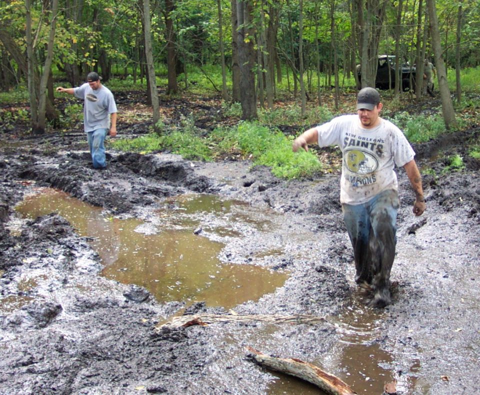 Muddy forest scene with two men wading through water and mud; a dark vehicle is visible in the background