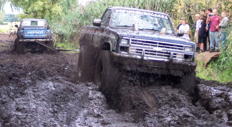 Front of a dark blue pickup truck struggling in deep mud, with another blue Chevrolet pickup truck being pulled out of the mud behind it