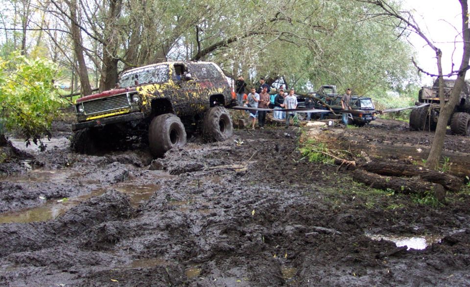 Muddy off-road vehicle with large tires stuck in deep mud; other vehicles and people visible in background