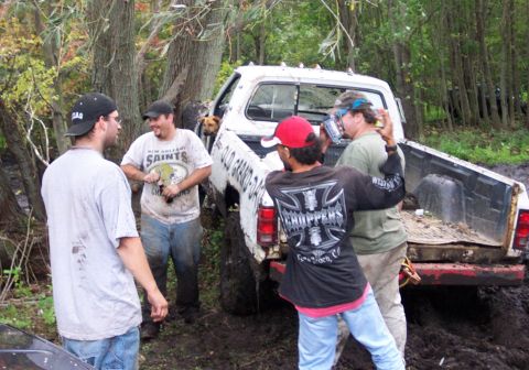 1978 Ford Bronco - Several people and a dog are near a white pickup truck that is stuck in a muddy, wooded area.