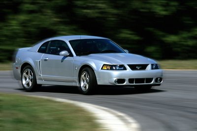 2003 Ford Mustang - Silver 2003 Ford Mustang coupe captured in motion on a paved road with blurred green foliage in the background.