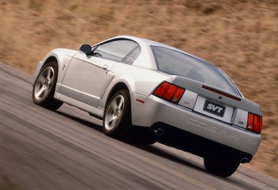 2003 Ford Mustang - A silver 2003 Ford SVT Mustang Cobra is captured in a rear 3/4 view, driving on a winding road.