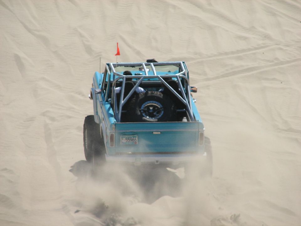 Blue caged early Bronco driving away kicking up sand on the dunes