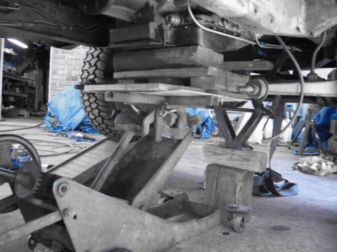 1978 Ford Bronco - Selective-color garage shot looking under a truck chassis with a leaf-spring axle supported on a floor jack stacked with wood blocks and a…