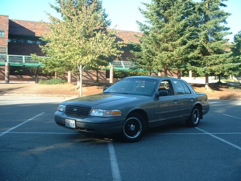 2000 Ford Crown Victoria - A gray Ford Crown Victoria is parked in a lot with a brick building and trees in the background.