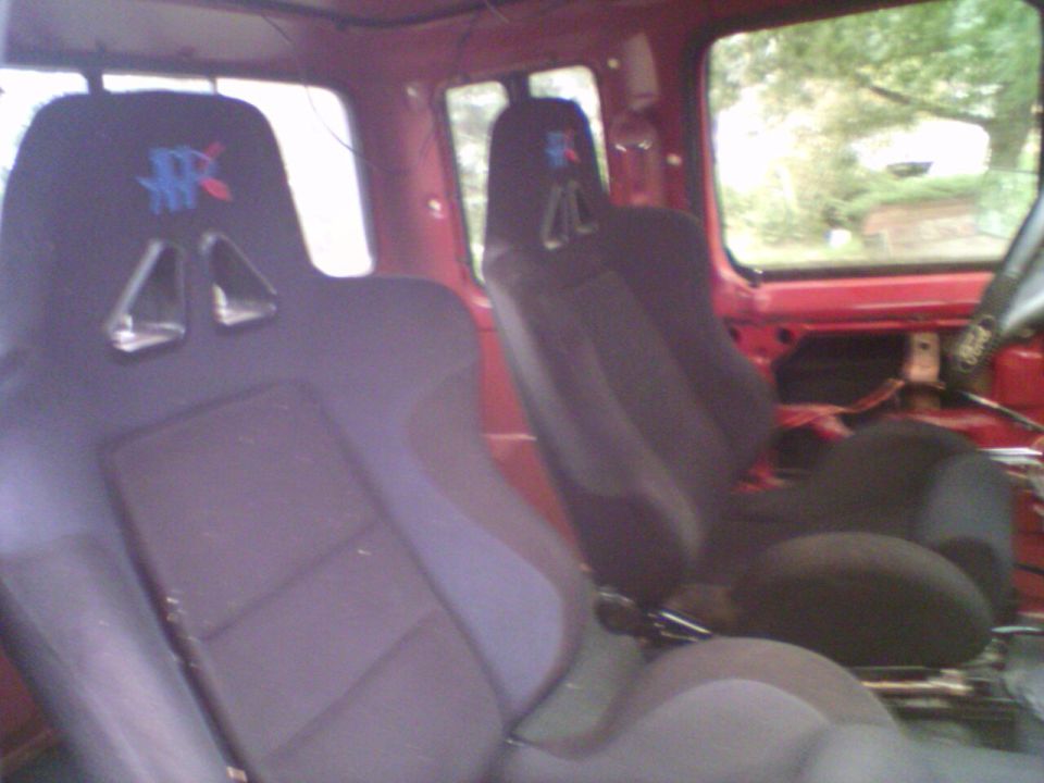 Gray bucket seats mounted in a stripped bare-metal red truck cab with exposed floor and seat brackets
