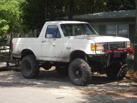 1989 Ford Bronco - A white, lifted pickup truck with off-road tires and a custom bumper is parked in a dirt lot.
