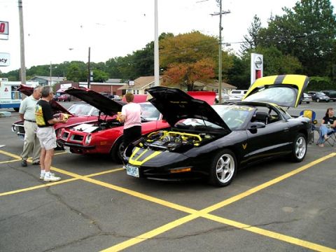 1994 Pontiac Trans Am - A black 1994 Pontiac Trans Am with yellow racing stripes is displayed with its hood and trunk open, alongside other vehicles.