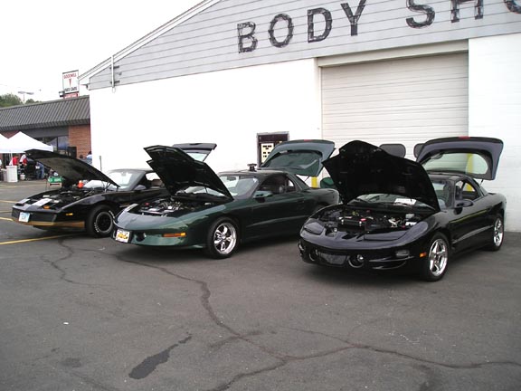 Three Pontiac Firebirds parked outside a body shop, hoods and trunks open