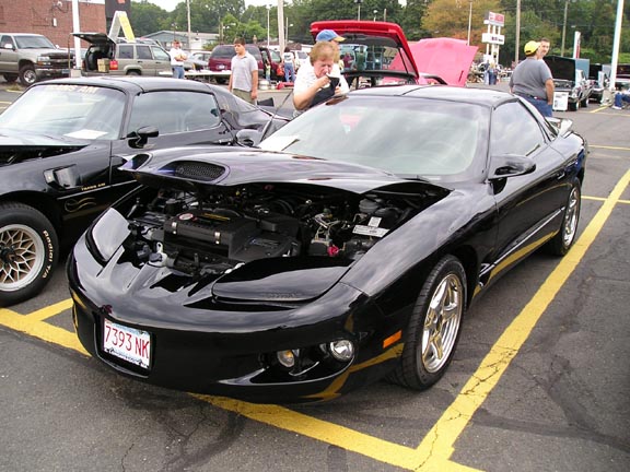 Black Pontiac Firebird Trans Am, hood open, showing engine. Another Trans Am is to the left. License plate 7393 NK