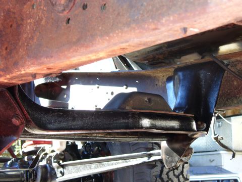 1978 Ford Bronco - Underbody view looking up at a black boxed frame rail and crossmember of a lifted 4x4, with a surface-rusted body floor above, a steering d…