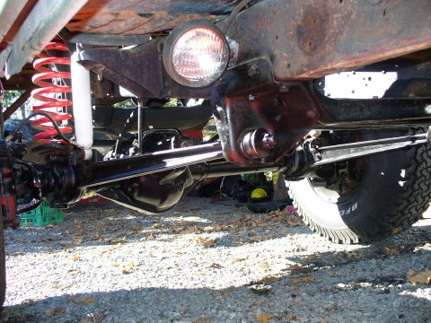 1978 Ford Bronco - Under-vehicle view along the front axle of a lifted truck, showing the drag link and tie rod, pitman arm, red coil springs, a white-bodied…