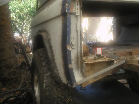 1966 Ford Bronco - Bare-metal and primer rear quarter panel of a truck undergoing bodywork, with tools and parts in a cluttered outdoor workshop.