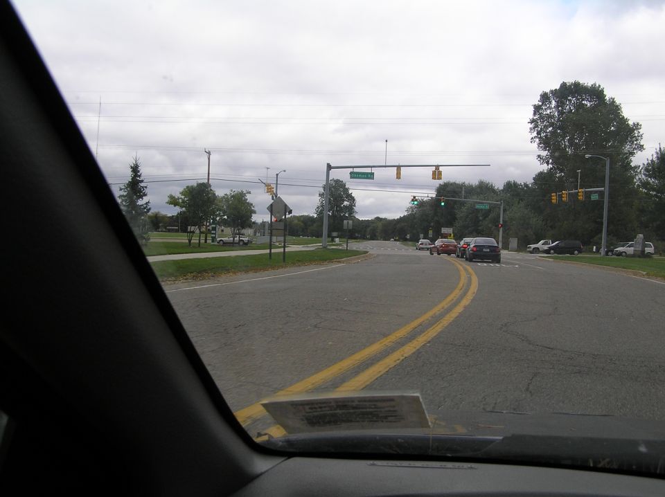 View from inside a car looking at a road intersection with several cars, trees, and traffic lights under a cloudy sky