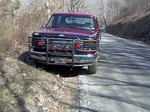 1994 Ford Bronco - A red Ford Bronco is parked on the side of a road with trees and brush in the background.