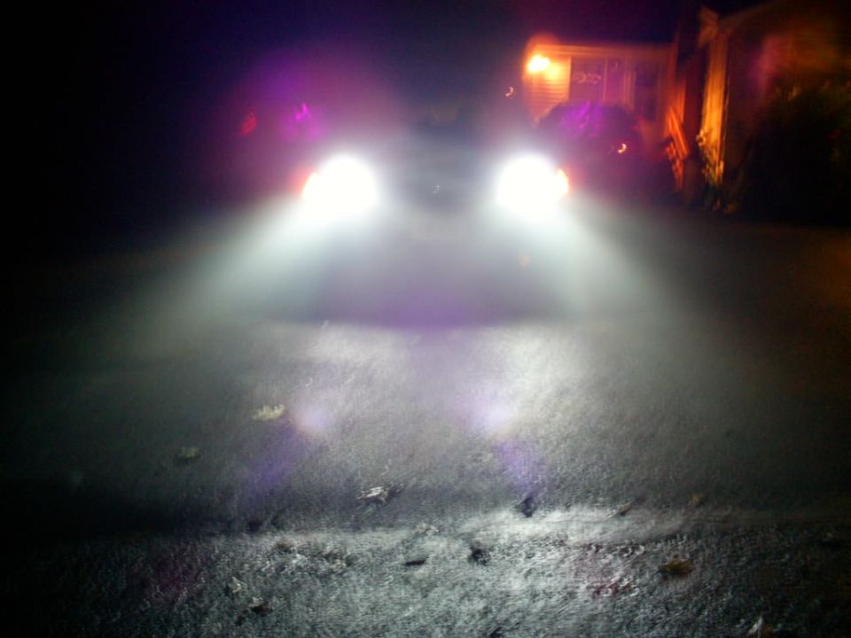 Car headlights illuminating a dark, possibly snowy, road at night; house lights visible in background