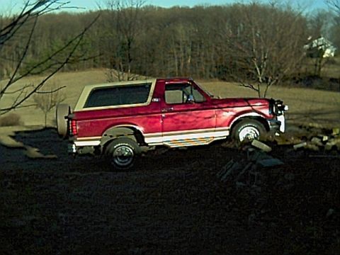 1994 Ford Bronco - This red Ford Bronco is parked on a dirt hill with trees and a house in the distance.