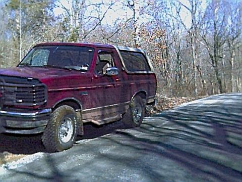 1994 Ford Bronco - A red Ford Bronco is parked on the side of a road with trees in the background.