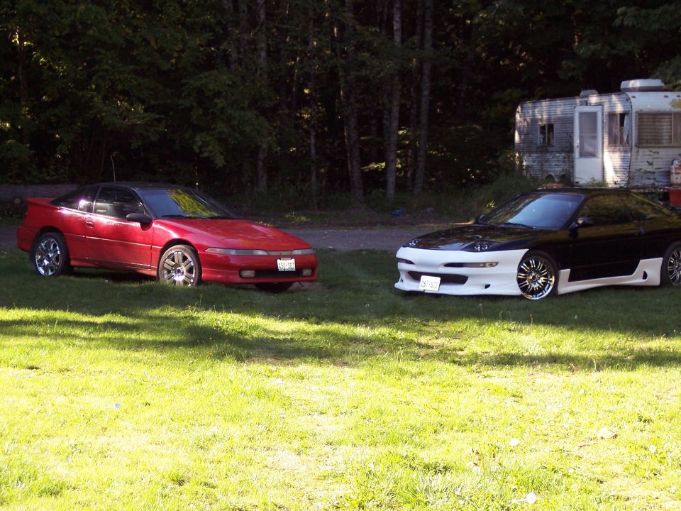Red and black/white sports cars parked on grass with a trailer in the background