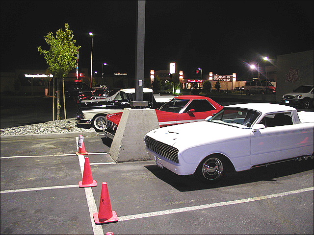 White early-60s compact car-pickup at night cruise-in with red coupe and shopping center behind