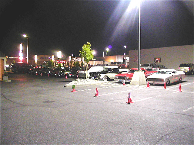 Classic cars parked at a nighttime cruise-in under parking lot lights
