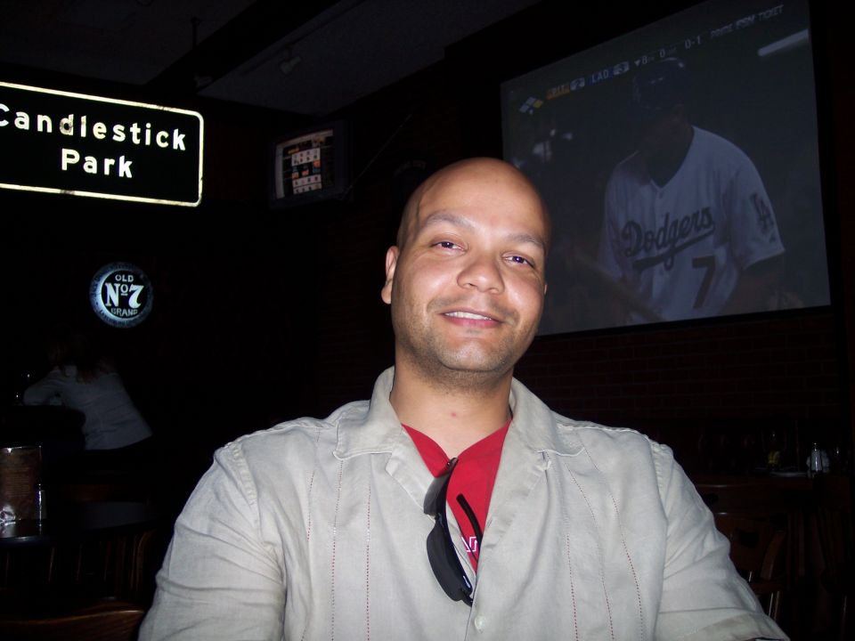 Man wearing a light colored shirt over a red shirt, with sunglasses hanging from his collar. A sign for Candlestick Park is visible