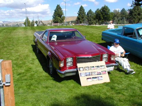1967 Ford Ranchero - A red 1977 Ford Ranchero is displayed at an outdoor event, with a board showing various car images and a blue pickup truck nearby.