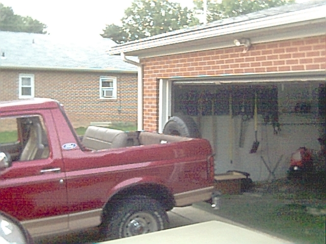 Red 1994 Ford Bronco with top removed, parked next to a garage