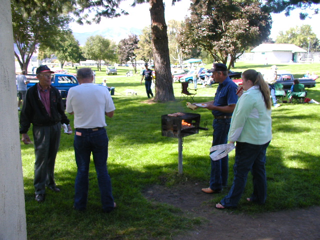 People around a park charcoal grill at a car club picnic, blue vintage pickup behind