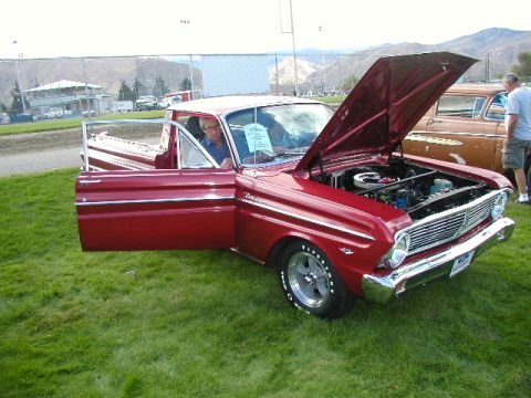 1967 Ford Ranchero - A man sits in a red pickup truck with its hood open, parked on grass at an outdoor event.