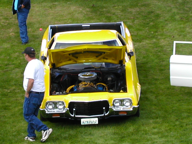 Yellow early-1970s Ford Ranchero with hood up showing V8 at a grassy car show