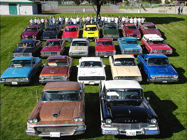 Overhead club photo of about twenty classic cars in rows on grass with owners behind