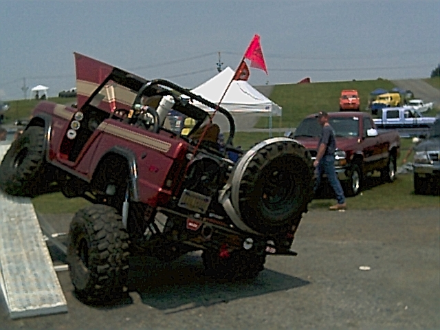 Red flatfender Jeep tilted nose-up on a teeter-totter obstacle with flag, at an off-road event