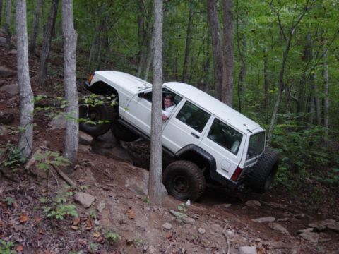 1991 Ford Bronco - A white Jeep Cherokee XJ navigates a steep, rocky incline in a wooded area.